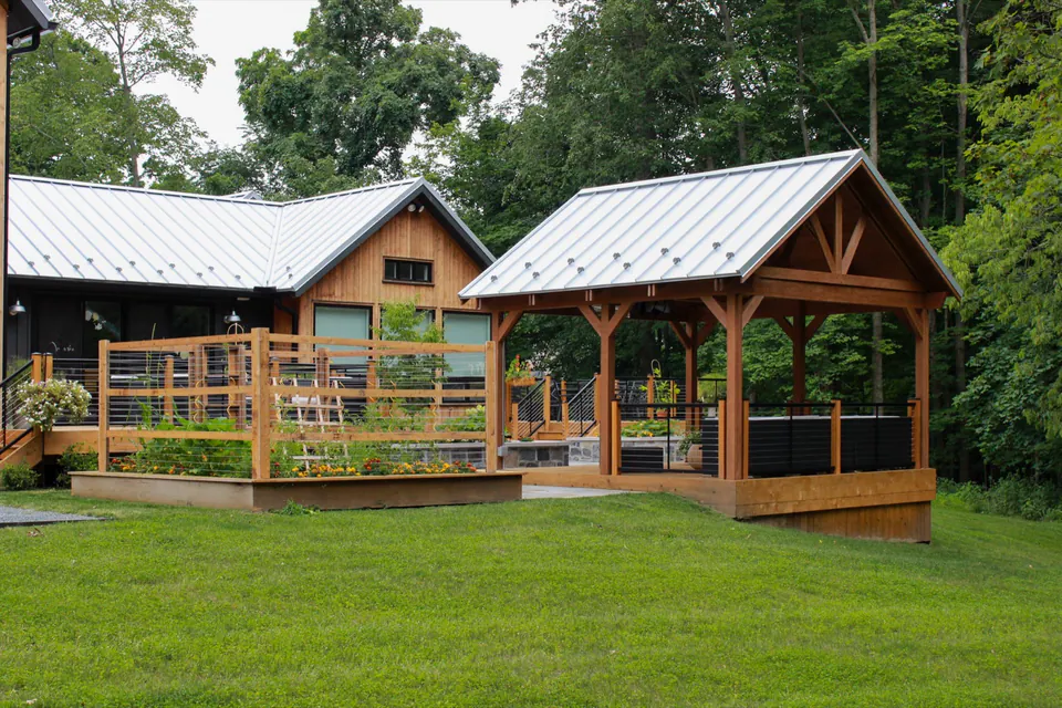 Covered wood pavilion and raised deck seen from lawn with cable railings, house, and wooded backdrop.